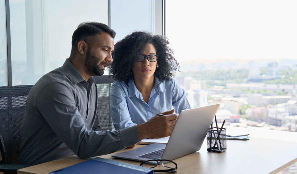 A man and woman in an office read from a laptop.