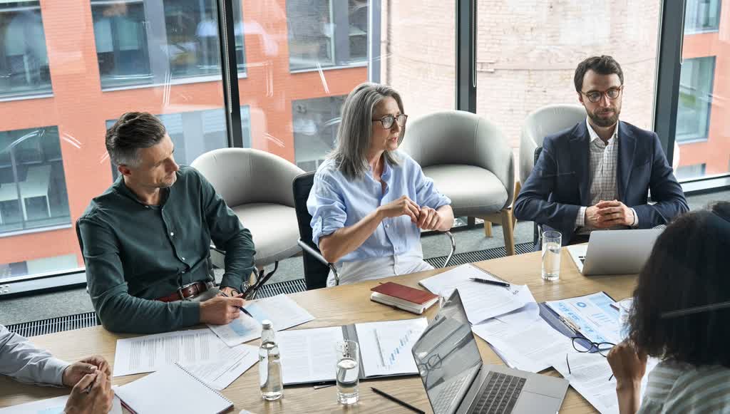 A group of people collaborate in a conference room with pens, papers, and laptops at the ready.